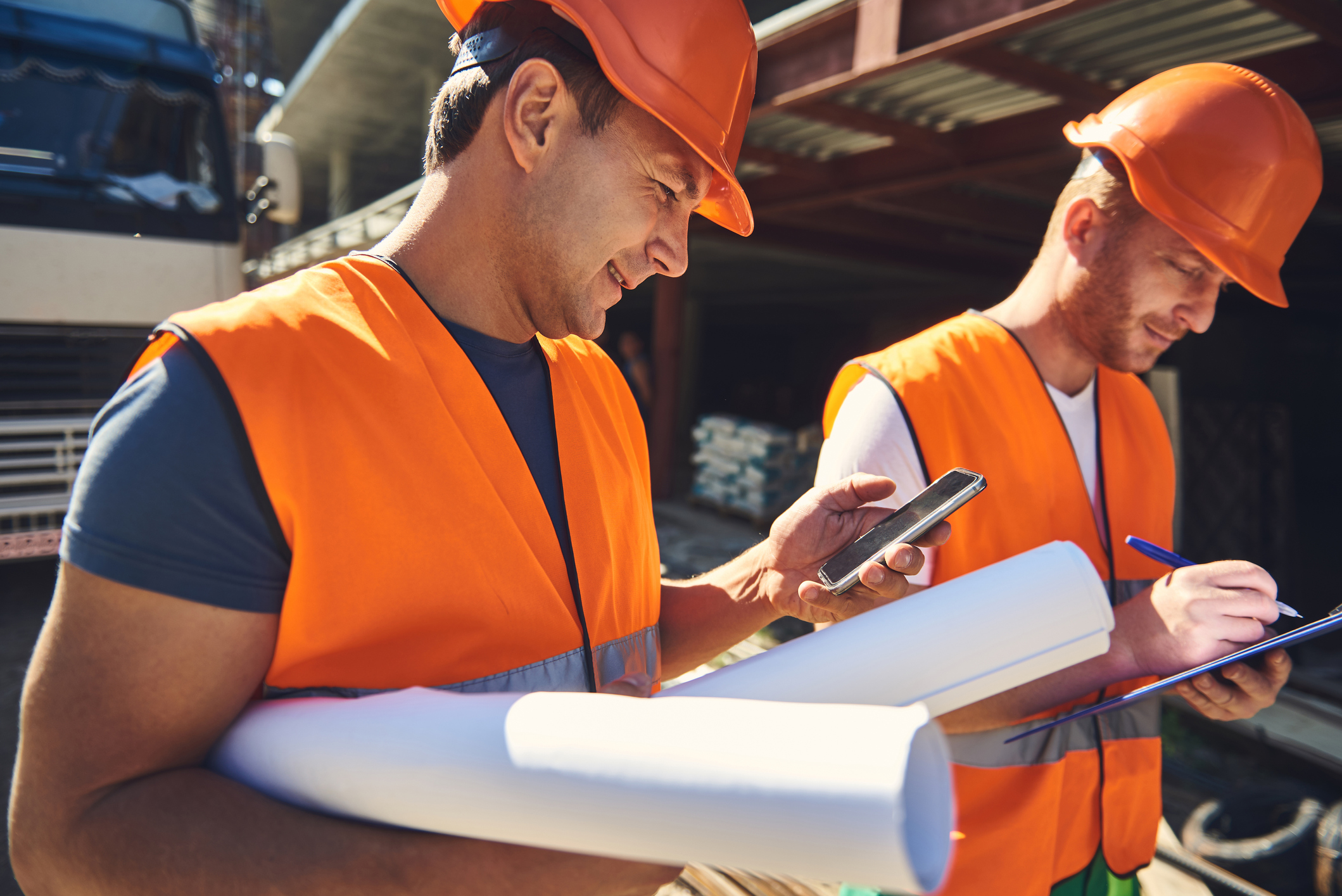Two workers are walking near construction site Waist up of two employees standing near the building while one of them holding smartphone