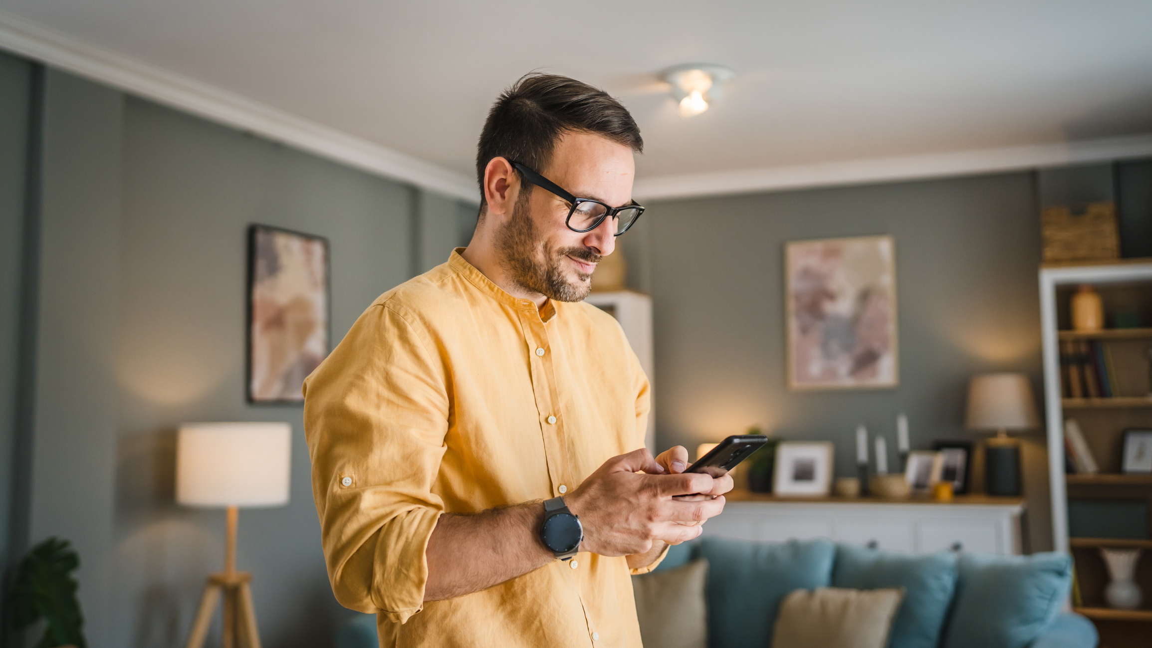 One man adult caucasian male with beard and eyeglasses stand at home happy smile use mobile phone smartphone to send sms text messages or browse internet online copy space