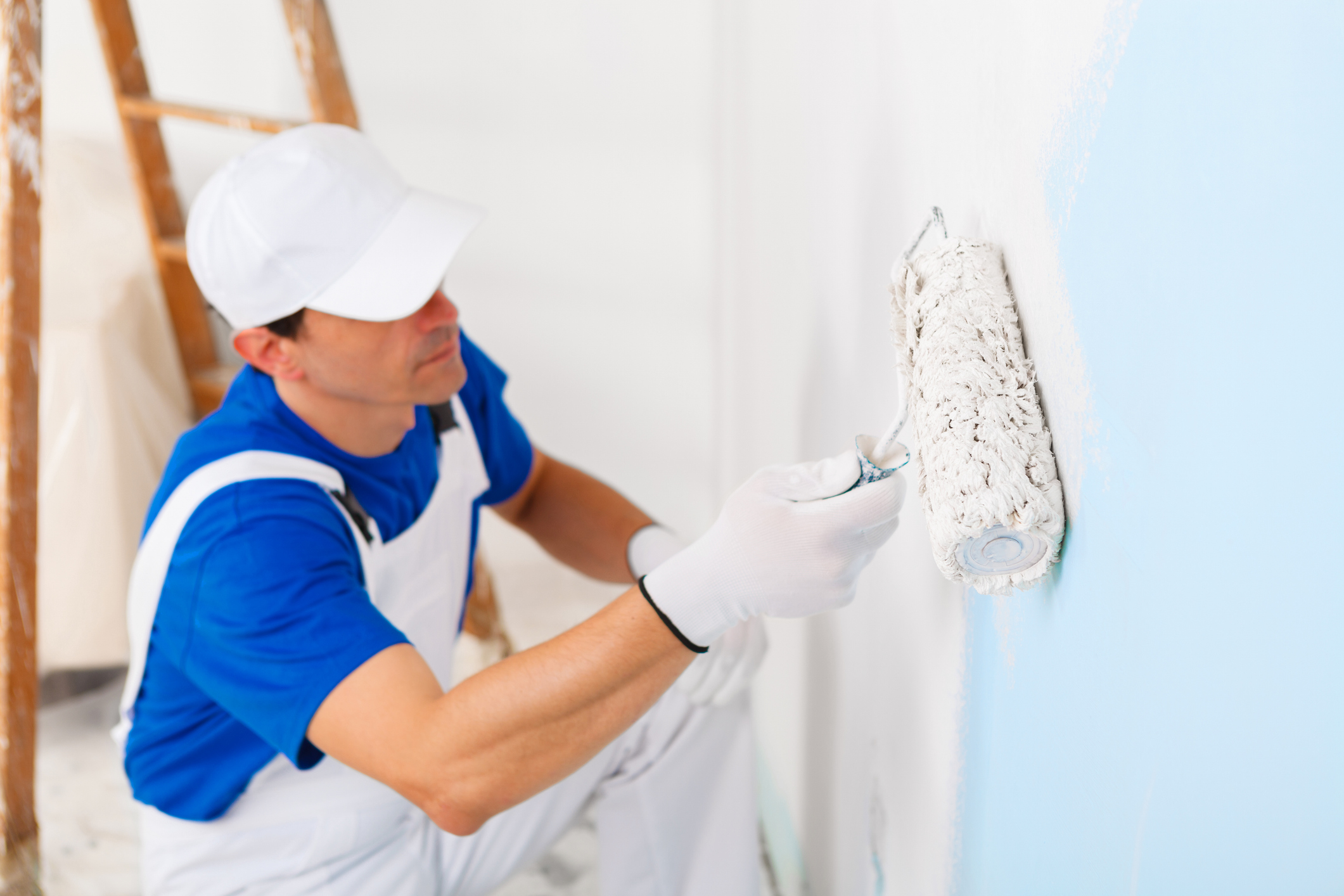side view of  painter in white dungarees, cap and gloves painting a wall with paint roller and wooden vintage ladder, selective focus on paint roller