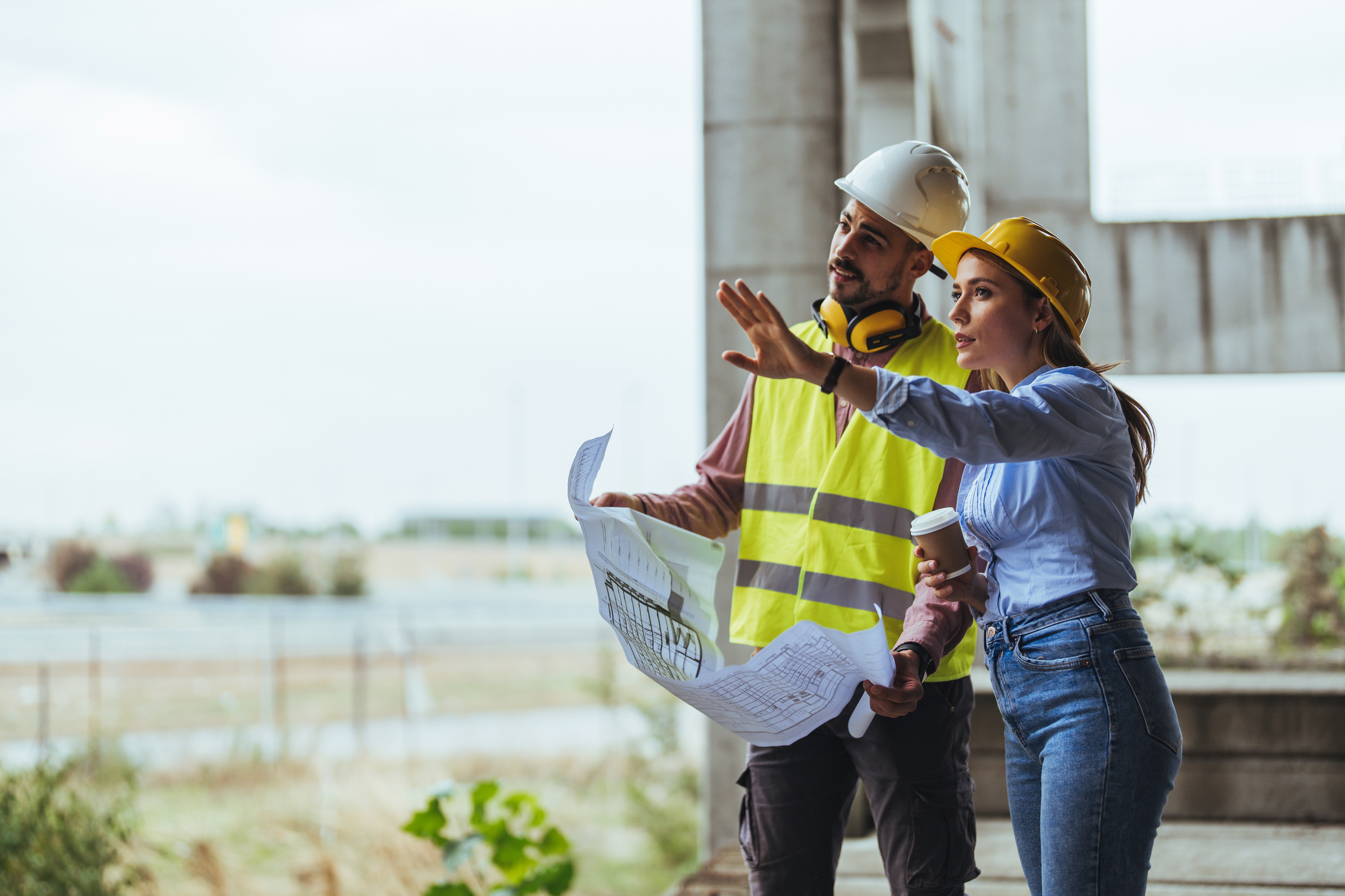 Young woman architect explaining blueprint to supervisor wearing safety vest at construction site. Mid adult contractor holding blueprint and understanding manager vision at construction site.